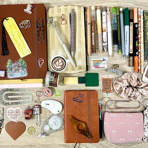 May include: A collection of stationery items including two brown notebooks, various pens, markers, and pencils, a striped pencil case, a pink pouch, a scrunchie, bookmarks, stickers, paper clips, a heart-shaped keychain, and a small white heart-shaped object.