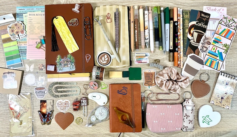 May include: A collection of stationery items including two brown notebooks, various pens, markers, and pencils, a striped pencil case, a pink pouch, a scrunchie, bookmarks, stickers, paper clips, a heart-shaped keychain, and a small white heart-shaped object.