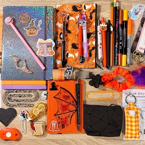 May include: A collection of stationery items including a holographic notebook, an orange bat-themed journal, various pens, pencils, markers, stickers, paper clips, sticky notes, book review cards, a small black pouch, and a bright orange scrunchie.