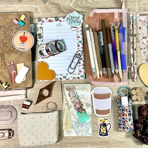 May include: A collection of stationery items including a brown notebook with a tree design, a notepad with the text Good Morning, a pink pencil case with pens, various stickers, bookmarks, paper clips, a small pouch, and a brown scrunchie.