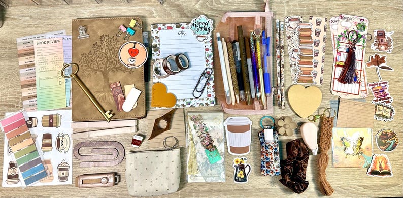 May include: A collection of stationery items including a brown notebook with a tree design, a notepad with the text Good Morning, a pink pencil case with pens, various stickers, bookmarks, paper clips, a small pouch, and a brown scrunchie.