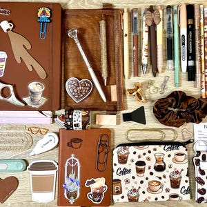 Puede incluir: Una colección de artículos de papelería que incluye dos cuadernos marrones, un estuche con cremallera de temática cafetera, varios bolígrafos, rotuladores, pegatinas, marcapáginas, listas de lectura, un coletero marrón y un llavero de cuentas.