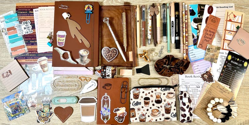 May include: A collection of stationery items including two brown notebooks, a coffee-themed zipped pouch, various pens, markers, stickers, bookmarks, reading lists, a brown scrunchie, and a beaded keychain, all featuring coffee and book motifs.