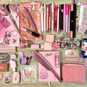 Pu&ograve; includere: Una collezione di articoli di cancelleria nei toni del rosa che include un quaderno a spirale, una penna a piuma, varie penne gel, pennarelli, foglietti adesivi, graffette, un piccolo astuccio a pois, adesivi decorativi e schede di lettura.