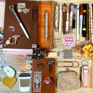 May include: A collection of stationery items including two brown notebooks, various pens, markers, paper clips, stickers, a yellow scrunchie, a small fuzzy coin purse, and reading list cards. The items feature coffee-themed graphics and text.