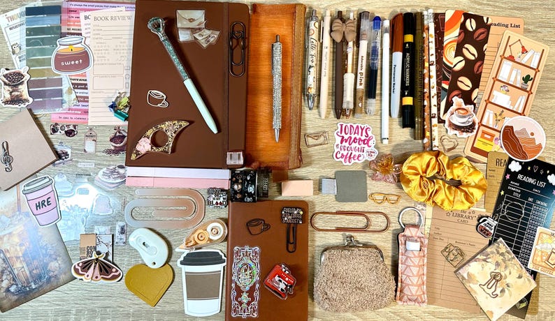 May include: A collection of stationery items including two brown notebooks, various pens, markers, paper clips, stickers, a yellow scrunchie, a small fuzzy coin purse, and reading list cards. The items feature coffee-themed graphics and text.