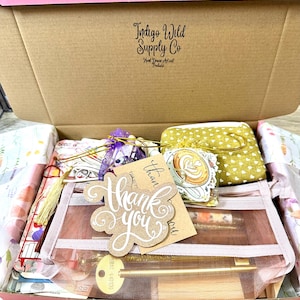 May include: A cardboard shipping box labelled Indigo Wild Supply Co containing a mesh pouch, a yellow heart-patterned fabric pouch, a purple organza bag, a gold key-shaped pen, and a brown paper tag with the handwritten text Thank you.