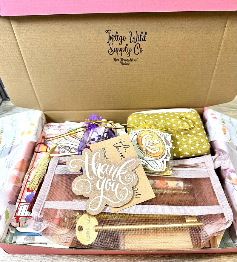 May include: A cardboard shipping box labelled Indigo Wild Supply Co containing a mesh pouch, a yellow heart-patterned fabric pouch, a purple organza bag, a gold key-shaped pen, and a brown paper tag with the handwritten text Thank you.