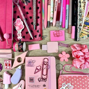 Pu&ograve; includere: Una collezione di articoli di cancelleria rosa che include due quaderni, una clip a forma di cuore, un astuccio con cuori neri, varie penne, pennarelli, adesivi, un elastico per capelli rosa, graffette, un piccolo taglierino e una pochette rosa con cuori bianchi.