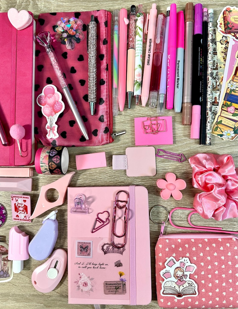 Pu&ograve; includere: Una collezione di articoli di cancelleria rosa che include due quaderni, una clip a forma di cuore, un astuccio con cuori neri, varie penne, pennarelli, adesivi, un elastico per capelli rosa, graffette, un piccolo taglierino e una pochette rosa con cuori bianchi.