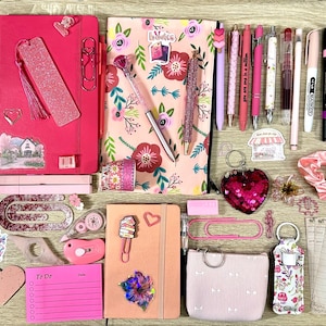 Pu&ograve; includere: Una collezione di articoli di cancelleria che include quaderni rosa e floreali, varie penne, foglietti adesivi, liste di lettura, graffette, un portachiavi a forma di cuore, una piccola custodia e adesivi decorativi in una disposizione piatta.