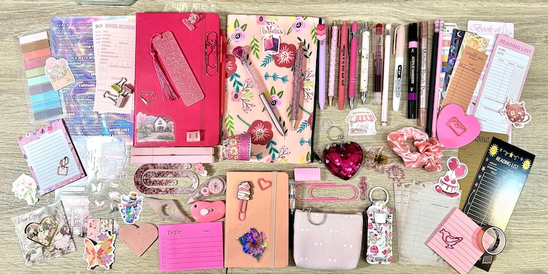 Pu&ograve; includere: Una collezione di articoli di cancelleria che include quaderni rosa e floreali, varie penne, foglietti adesivi, liste di lettura, graffette, un portachiavi a forma di cuore, una piccola custodia e adesivi decorativi in una disposizione piatta.