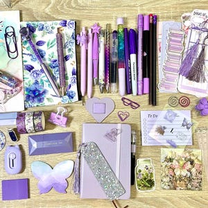 May include: A collection of purple and lavender stationery items including a spiral notebook, a bound journal, various pens, pencils, sticky notes, bookmarks, a scrunchie, and decorative stickers featuring floral, butterfly, and book-themed designs.