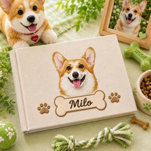 May include: A rectangular photo album with a light beige fabric cover featuring an embroidered portrait of a Corgi dog. Below the dog is an embroidered bone shape containing the name Milo in brown script, flanked by two small brown paw prints.