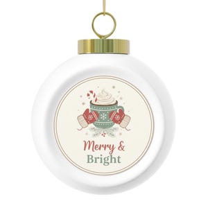 Może przedstawiać: Biała bombka choinkowa ze złotą nasadką i wieszakiem. Ozdoba ma okrągły wzór z kubkiem gorącego kakao, rękawicami i napisem "Merry & Bright" w kolorze czerwonym i zielonym. Świąteczna dekoracja.