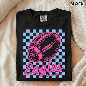 K&ouml;nnte beinhalten: Ein schwarzes T-Shirt mit einer Grafik eines Footballs, zentriert &uuml;ber einem hellblau-schwarzen Schachbrettmuster. Unter dem Football steht das Wort Eagles in einer rosa Schreibschrift, gefolgt vom Text School Name in kleinerer Schrift.