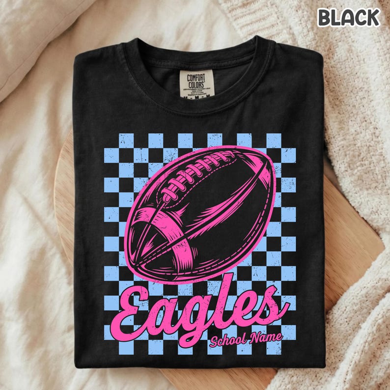 K&ouml;nnte beinhalten: Ein schwarzes T-Shirt mit einer Grafik eines Footballs, zentriert &uuml;ber einem hellblau-schwarzen Schachbrettmuster. Unter dem Football steht das Wort Eagles in einer rosa Schreibschrift, gefolgt vom Text School Name in kleinerer Schrift.