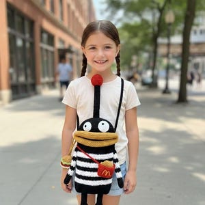 以下が含まれることがあります： 手作りのかぎ針編みのクロスボディバッグ。白と黒のストライプのボディ、黄色の口、赤い財布が特徴の漫画のキャラクターの形をしています。バッグには黒いストラップと小さなハンバーガーのキーホルダーが付いています。