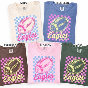 K&ouml;nnte beinhalten: F&uuml;nf kurz&auml;rmelige T-Shirts in den Farben Elfenbein, Jeansblau, Espresso, Bl&uuml;tenrosa und Moosgr&uuml;n. Jedes Shirt zeigt eine Grafik eines Footballs &uuml;ber einem Karomuster mit dem Text "Eagles" und "School Name" in einer Schreibschrift.