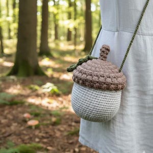 May include: A crocheted acorn-shaped crossbody bag with a light brown top and a light gray base. The bag has a green strap and is set against a blurred forest background.