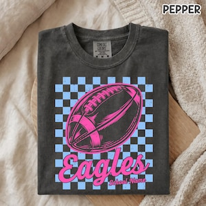 K&ouml;nnte beinhalten: Ein anthrazitfarbenes T-Shirt mit einem hellblau-schwarzen Karomuster im Hintergrund. Zentriert ist eine leuchtend rosa Grafik eines Footballs abgebildet, darunter steht das Wort Eagles in kursiver rosa Schrift und School Name in kleiner.