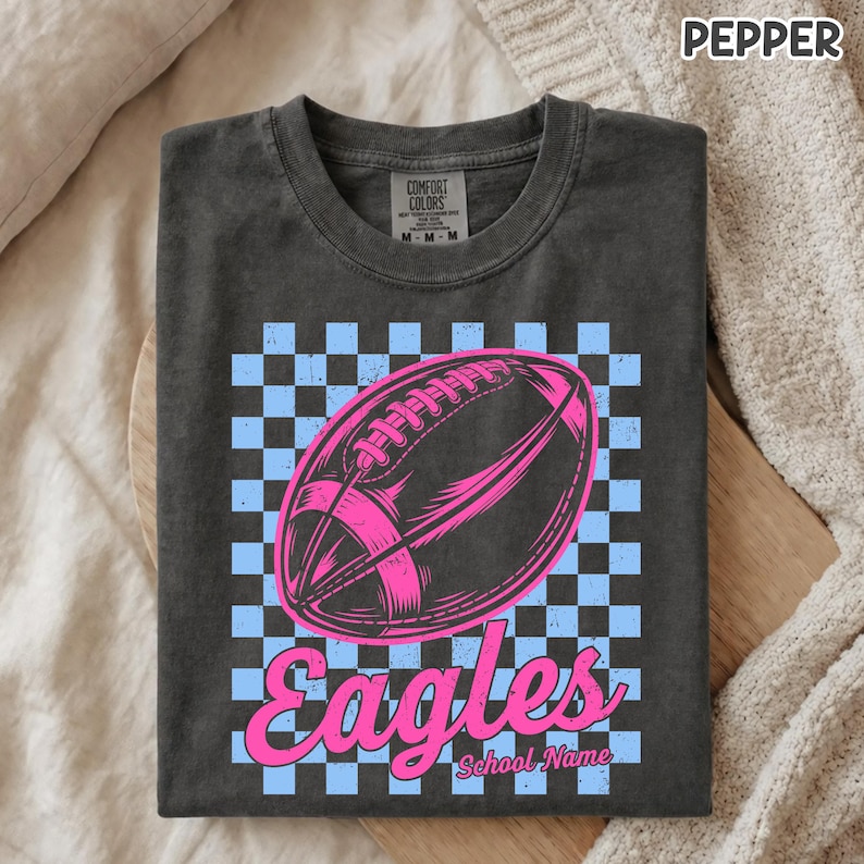 K&ouml;nnte beinhalten: Ein anthrazitfarbenes T-Shirt mit einem hellblau-schwarzen Karomuster im Hintergrund. Zentriert ist eine leuchtend rosa Grafik eines Footballs abgebildet, darunter steht das Wort Eagles in kursiver rosa Schrift und School Name in kleiner.