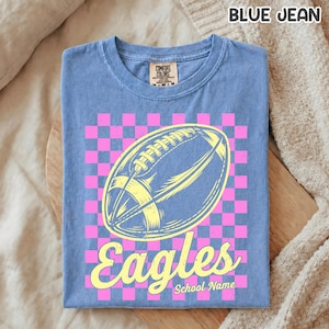K&ouml;nnte beinhalten: Ein blaues T-Shirt in Jeansoptik mit einer Grafik eines gelben Footballs, der &uuml;ber einem rosa-blauen Karomuster zentriert ist. Das Wort Eagles ist in gelber Schreibschrift unter dem Football gedruckt, darunter steht der Text School Name.