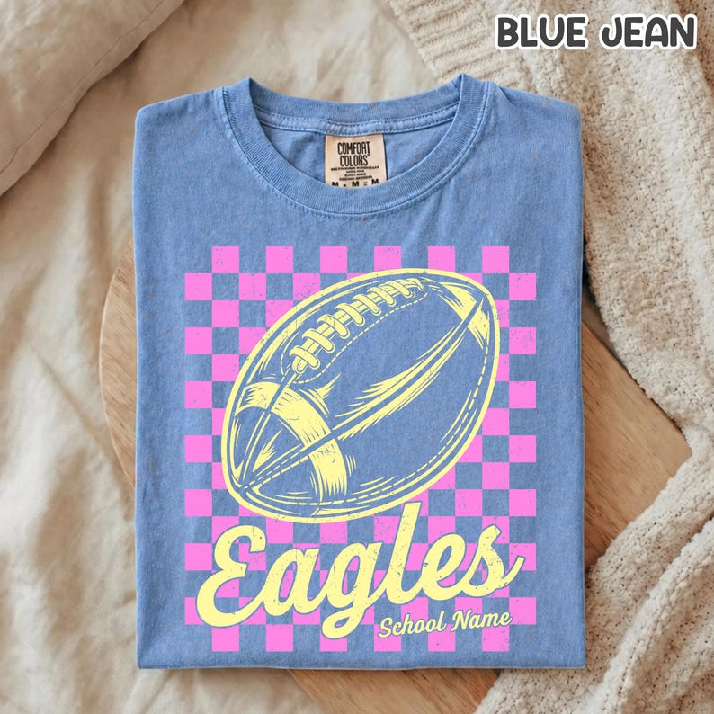 K&ouml;nnte beinhalten: Ein blaues T-Shirt in Jeansoptik mit einer Grafik eines gelben Footballs, der &uuml;ber einem rosa-blauen Karomuster zentriert ist. Das Wort Eagles ist in gelber Schreibschrift unter dem Football gedruckt, darunter steht der Text School Name.