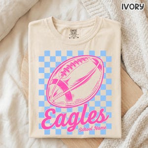 K&ouml;nnte beinhalten: Ein elfenbeinfarbenes T-Shirt mit einer Grafik eines pinkfarbenen Footballs, zentriert &uuml;ber einem hellblauen und elfenbeinfarbenen Schachbrettmuster. Unter dem Football steht das Wort "Eagles" in pinker Schreibschrift und "School Name" in Pink.