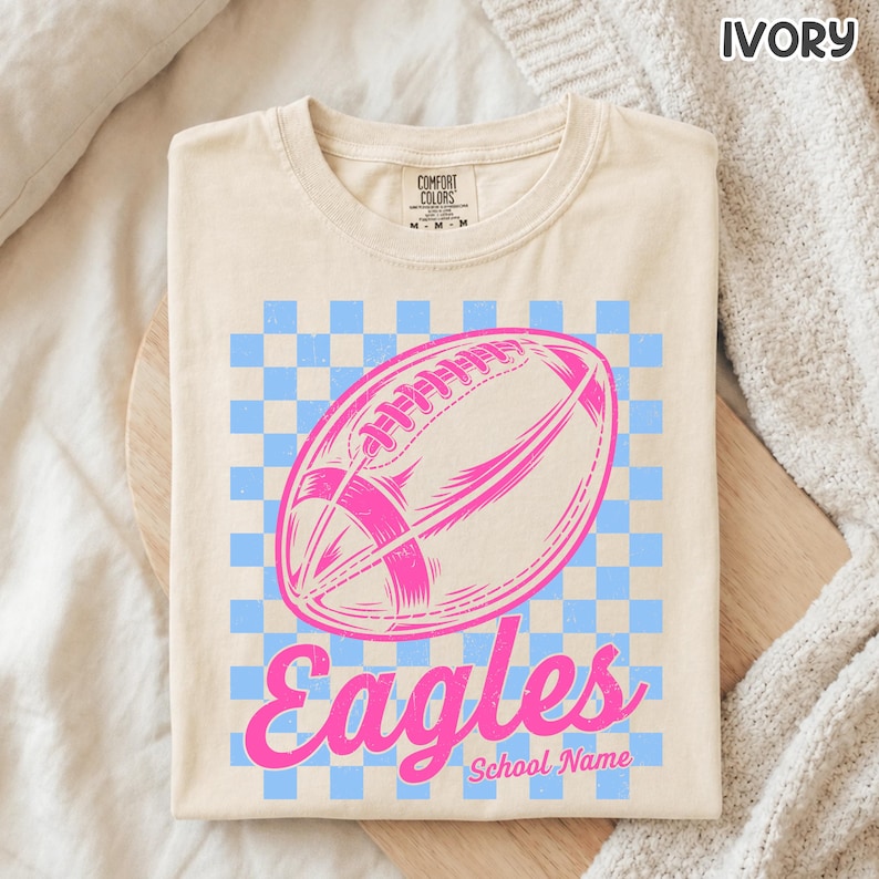 K&ouml;nnte beinhalten: Ein elfenbeinfarbenes T-Shirt mit einer Grafik eines pinkfarbenen Footballs, zentriert &uuml;ber einem hellblauen und elfenbeinfarbenen Schachbrettmuster. Unter dem Football steht das Wort "Eagles" in pinker Schreibschrift und "School Name" in Pink.