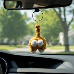 Op de afbeelding: Een handgemaakte, gehaakte kattenkont auto-hanger. De hanger is voornamelijk mosterdgeel met witte accenten en bruine strepen. Hij hangt aan een goudkleurige ring, bevestigd aan de achteruitkijkspiegel van een auto. De achtergrond is wazig.