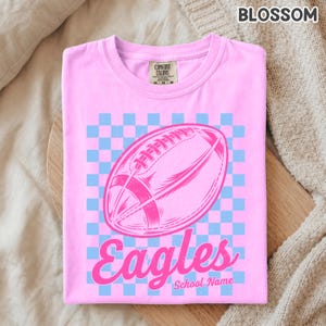 K&ouml;nnte beinhalten: Ein hellrosa T-Shirt mit kurzen &Auml;rmeln und einem hellblau-rosa karierten Hintergrund. In der Mitte befindet sich eine rosa Grafik eines American Footballs mit dem Wort Eagles in Schreibschrift und dem Text School Name darunter.
