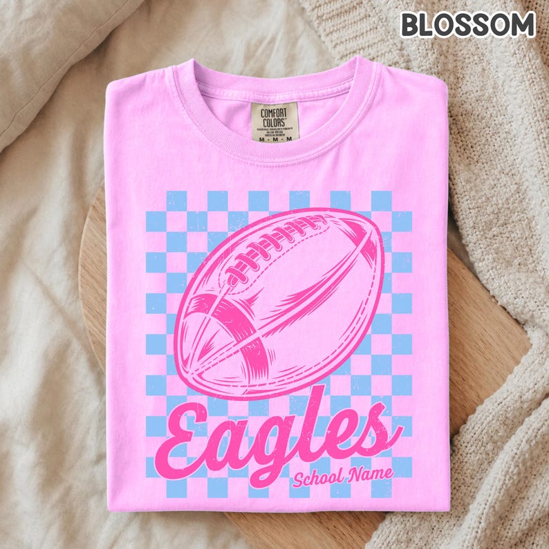 K&ouml;nnte beinhalten: Ein hellrosa T-Shirt mit kurzen &Auml;rmeln und einem hellblau-rosa karierten Hintergrund. In der Mitte befindet sich eine rosa Grafik eines American Footballs mit dem Wort Eagles in Schreibschrift und dem Text School Name darunter.