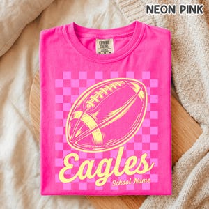 K&ouml;nnte beinhalten: Ein neonpinkes T-Shirt mit Rundhalsausschnitt und einer gelben Grafik eines American Footballs auf einem hellrosa Schachbrettmuster. Das Wort Eagles ist in gelber Schreibschrift unter dem Ball gedruckt, mit dem Text School Name darunter.