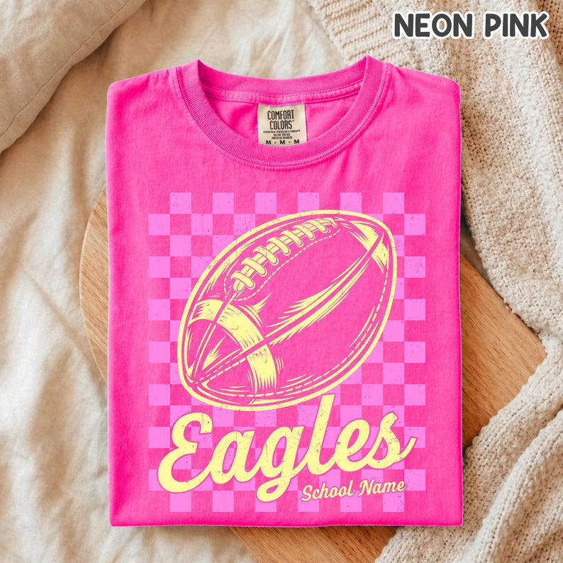 K&ouml;nnte beinhalten: Ein neonpinkes T-Shirt mit Rundhalsausschnitt und einer gelben Grafik eines American Footballs auf einem hellrosa Schachbrettmuster. Das Wort Eagles ist in gelber Schreibschrift unter dem Ball gedruckt, mit dem Text School Name darunter.