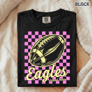 K&ouml;nnte beinhalten: Ein schwarzes T-Shirt mit einer Grafik eines gelben Footballs, zentriert &uuml;ber einem rosa-schwarzen Karomuster. Unter dem Football steht das Wort Eagles in einer gelben Schreibschrift, gefolgt vom Text School Name.