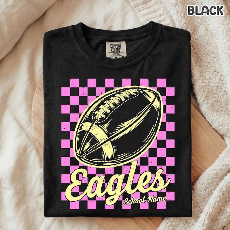 K&ouml;nnte beinhalten: Ein schwarzes T-Shirt mit einer Grafik eines gelben Footballs, zentriert &uuml;ber einem rosa-schwarzen Karomuster. Unter dem Football steht das Wort Eagles in einer gelben Schreibschrift, gefolgt vom Text School Name.