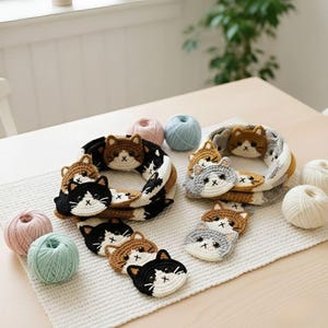 May include: Crocheted cat-themed items, including coasters and decorative rings. The coasters feature cat faces in black, brown, and gray. The rings are in matching colors with a repeating cat face pattern. Yarn balls in pink and blue are also present.