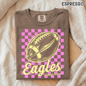 K&ouml;nnte beinhalten: Ein espressobraunes T-Shirt mit einer Grafik eines Footballs, zentriert &uuml;ber einem rosa-braunen Karomuster. Das Wort Eagles ist in gelber Schreibschrift unter dem Football gedruckt, mit dem Text School Name darunter.