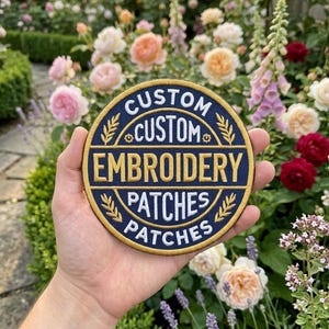 May include: A circular embroidered patch with a dark blue background and gold border. The text reads CUSTOM CUSTOM EMBROIDERY PATCHES PATCHES in white and gold lettering, arranged in concentric circles with small gold wheat-like accents.