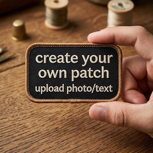 May include: A rectangular black embroidered patch with a brown stitched border. The patch features white text that reads: create your own patch upload photo/text. The design is centered on a black fabric background with clear, legible lettering.
