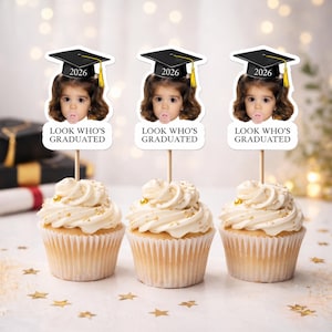 May include: Three cupcakes with white frosting and gold sprinkles. Each cupcake has a topper with a child's face wearing a graduation cap and the year 2026. The text on the topper reads "LOOK WHO'S GRADUATED".