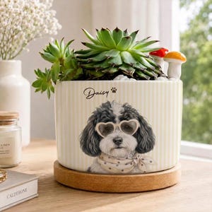 May include: A cylindrical ceramic planter with a cream and white vertical stripe pattern, featuring a graphic of a black and white dog wearing heart-shaped glasses and a polka dot scarf. The name Daisy and a paw print are printed above the dog.