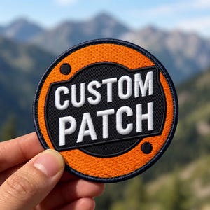 Peut inclure: Un écusson brodé circulaire avec une bordure orange vif et un centre noir. Le texte CUSTOM PATCH est brodé en lettres capitales blanches et grasses au milieu. L'écusson présente un contour bleu marine et deux petits cercles noirs.