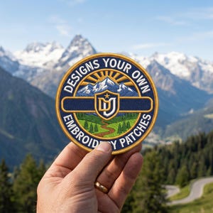 Peut inclure: Un écusson brodé circulaire avec une bordure bleue et dorée portant le texte DESIGNS YOUR OWN EMBROIDERY PATCHES. Le centre présente un paysage de montagne, une vallée verte avec un chemin et un écusson central avec les lettres DU et une étoile.