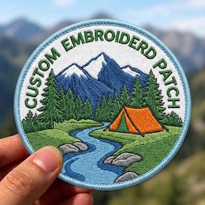 May include: A circular embroidered patch featuring a landscape scene with a blue river, green pine trees, a bright orange tent, and snow-capped mountains. The text CUSTOM EMBROIDRD PATCH is stitched in green along the top edge of the white border.