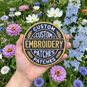 May include: A circular embroidered patch with a dark blue background and gold border. The text reads CUSTOM CUSTOM EMBROIDERY PATCHES PATCHES in white and gold lettering, accented by small gold wheat-like stalks arranged in a circular pattern.