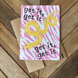 May include: A rectangular greeting card featuring a yellow snake illustration on a pink and white zebra-striped background. The black text reads get it, get it, get it, get it. The card is placed on top of a plain brown paper envelope.