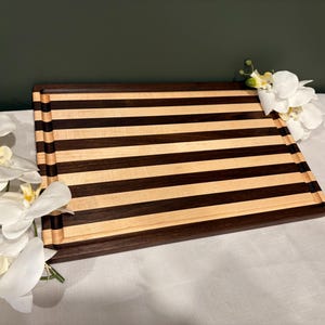 May include: A rectangular wooden chopping board with alternating dark brown and light wood stripes. The board has a dark brown border and a juice groove. White orchids are placed on the sides.