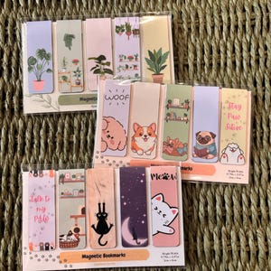 May include: Three sets of magnetic bookmarks in clear plastic packaging. The designs feature illustrations of potted plants, various dog breeds, cats, and text including "WOOF", "Stay Paw Sitive", "talk to my PAW", and "Meow". Each bookmark is 2cm x 6cm.